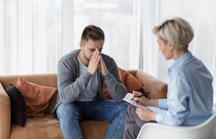 Smutny mężczyzna z złączonymi dłońmi podczas sesji terapeutycznej, psycholog robi notatki.