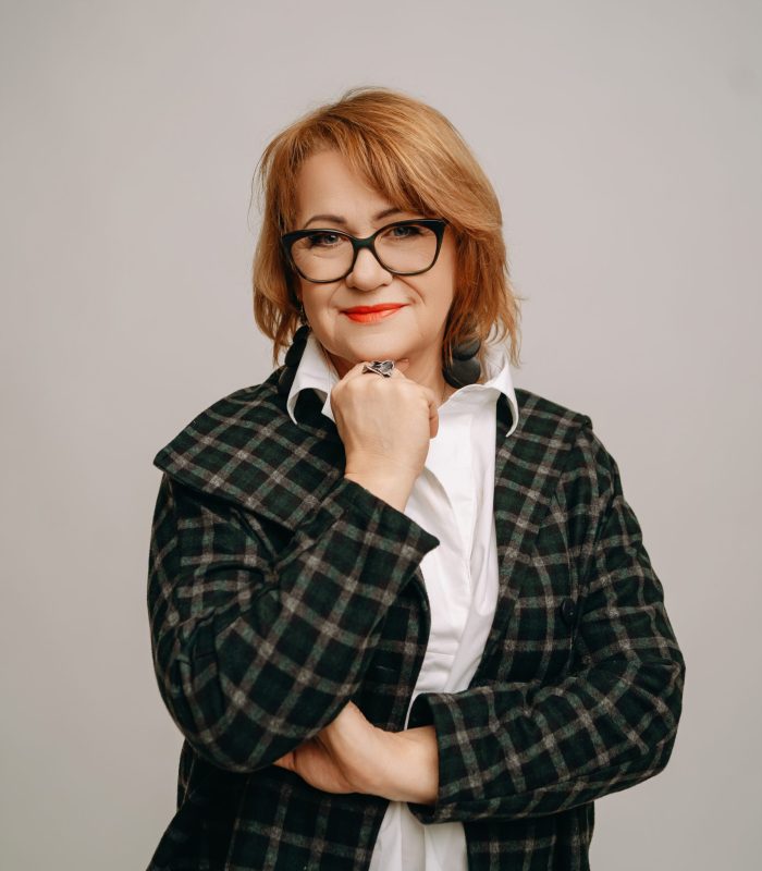 Portret uśmiechniętej kobiety w średnim wieku z rudymi włosami, ubranej w białą koszulę i luźną, kratkowaną, ciemnozieloną kurtkę. Kobieta ma na sobie czarne okulary i duży pierścień, a dłoń zgiętą w łokciu trzyma pod brodą. Tło jest jednolite, jasnoszare.